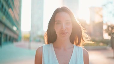 Çilli, koyu renk saçlı, beyaz bluzlu ve kot pantolonlu hoş bir genç kadın modern şehir arka planında gülümsüyor. Arkadaki ışıkta gülümseyen uzun kirpikli güzel kız.