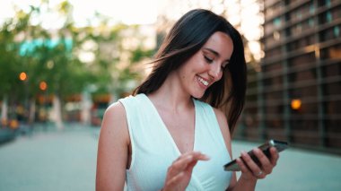 Çilli, siyah saçlı, beyaz bluzlu güzel bir kadın elinde akıllı telefonuyla sokakta yürüyor. Kız modern şehir arka planında cep telefonu kullanıyor