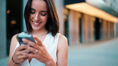 Çilli, siyah saçlı, beyaz bluzlu güzel bir kadın elinde akıllı telefonuyla sokakta yürüyor. Kız modern şehir arka planında cep telefonu kullanıyor