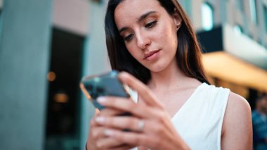 Çilli, siyah saçlı, beyaz bluzlu güzel bir kadın elinde akıllı telefonuyla sokakta yürüyor. Kız modern şehir arka planında cep telefonu kullanıyor