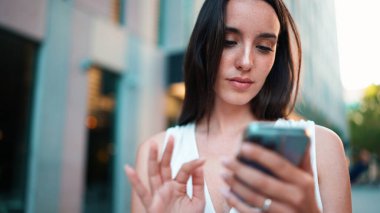 Çilli, siyah saçlı, beyaz bluzlu güzel bir kadın elinde akıllı telefonuyla sokakta yürüyor. Kız modern şehir arka planında cep telefonu kullanıyor