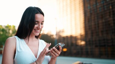 Çilli, siyah saçlı, beyaz bluzlu güzel bir kadın elinde akıllı telefonuyla sokakta yürüyor. Kız modern şehir arka planında cep telefonu kullanıyor