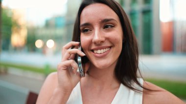 Çilli, koyu renk saçlı, beyaz bluzlu güzel bir kadın elinde telefonla bankta oturuyor. Cep telefonuyla konuşan sevimli kız modern şehir geçmişi.