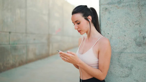 Young athletic woman with long ponytail wearing beige sports top in wired headphones, stands with mobile phone in her hands