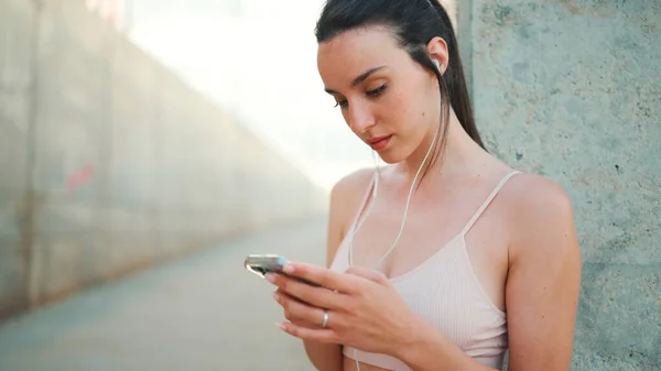 Young athletic woman with long ponytail wearing beige sports top in wired headphones, stands with mobile phone in her hands