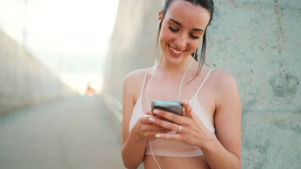 Young athletic woman with long ponytail wearing beige sports top in wired headphones, stands with mobile phone in her hands, listening to music and smiling