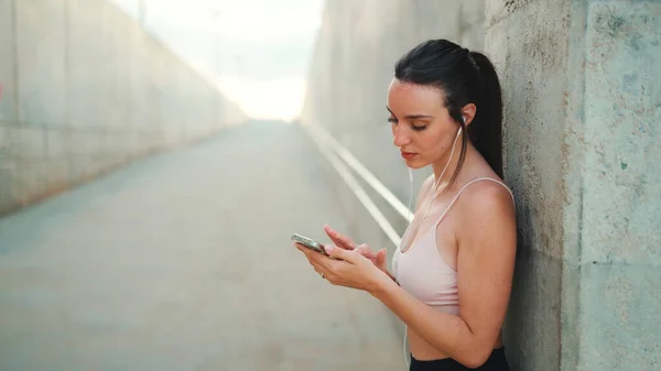Young athletic woman with long ponytail wearing beige sports top in wired headphones, stands with mobile phone in her hands