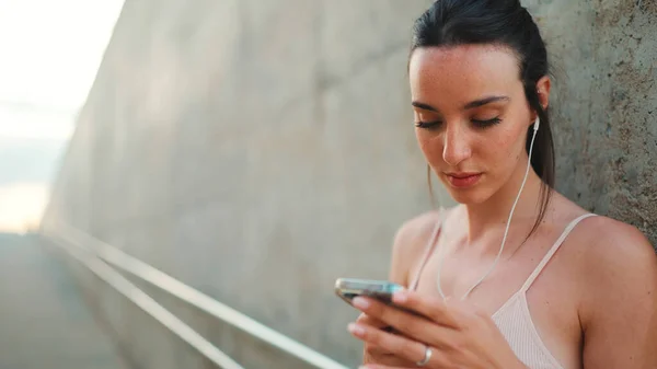 Young athletic woman with long ponytail wearing beige sports top in wired headphones, stands with mobile phone in her hands