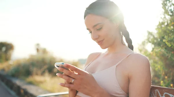 Young athletic woman with braided pigtail wearing beige sports top is sitting on bench with mobile phone in her hands, watching social networks, photos and videos