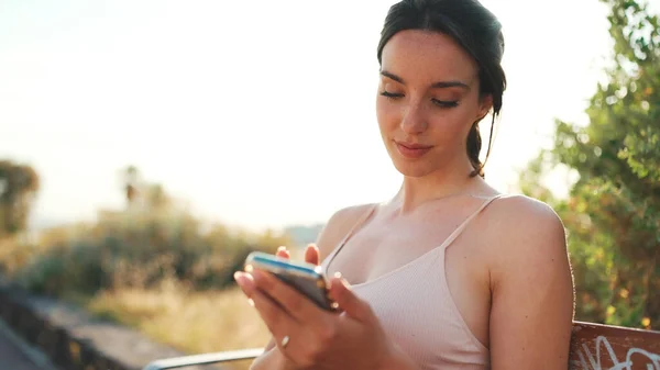 Young athletic woman with braided pigtail wearing beige sports top is sitting on bench with mobile phone in her hands, watching social networks, photos and videos