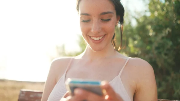 Young athletic woman with braided pigtail wearing beige sports top is sitting on bench with mobile phone in her hands, watching social networks, photos and videos