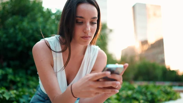 Cute young woman with freckles and dark loose hair wearing white top is using mobile phone. Beautiful girl sits in city park looking through photos, videos on mobile phone. Camera moves around