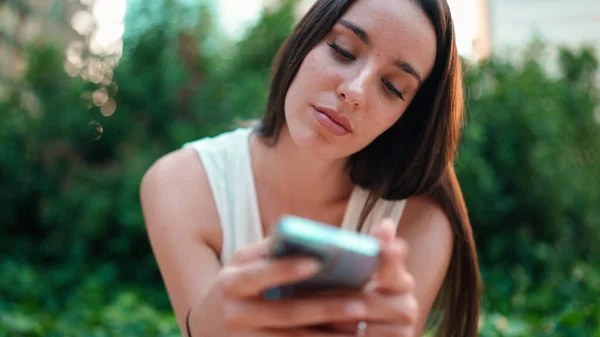 Cute young woman with freckles and dark loose hair wearing white top is using mobile phone. Beautiful girl sits in city park looking through photos, videos on mobile phone. Camera moves around