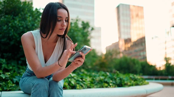 Cute young woman with freckles and dark loose hair wearing white top is using mobile phone. Beautiful girl sits in city park looking through photos, videos on mobile phone. Camera moves around