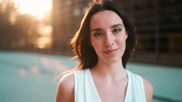 Close-up portrait of young woman with freckles and dark loose hair and long eyelashes wearing white top looking straight at the camera. Beautiful girl on modern city background