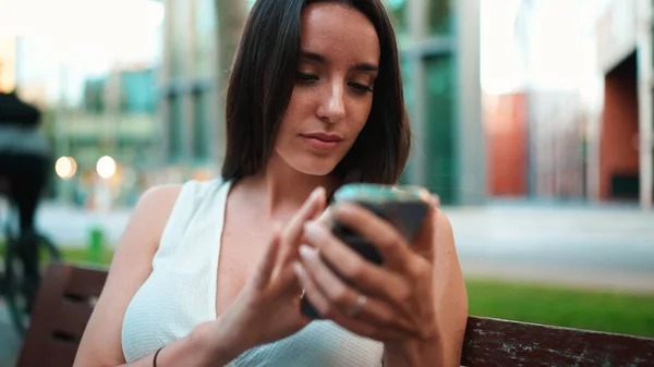 Beautiful woman with freckles and dark loose hair wearing white top sits on bench with phone in her hands. Cute smiling girl watching photo, video on mobile phone modern city background