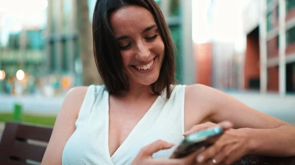 Beautiful woman with freckles and dark loose hair wearing white top sits on bench with phone in her hands. Cute smiling girl watching photo, video on mobile phone modern city background