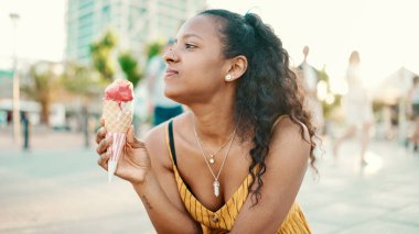 Kentin arka planında elinde dondurma olan uzun kıvırcık saçlı, gülümseyen bir kadının yakın plan portresi. Sıcak ve güneşli bir günde dondurma test eden mutlu bir kızın önden yakın çekimi.