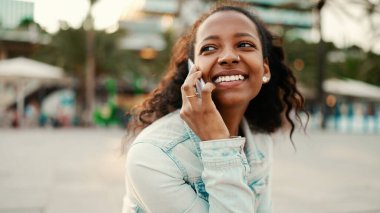 Kentin arka planında cep telefonu kullanan uzun kıvırcık saçlı genç bir kadın. Akıllı telefondan konuşan bir kızın yakın çekimi.