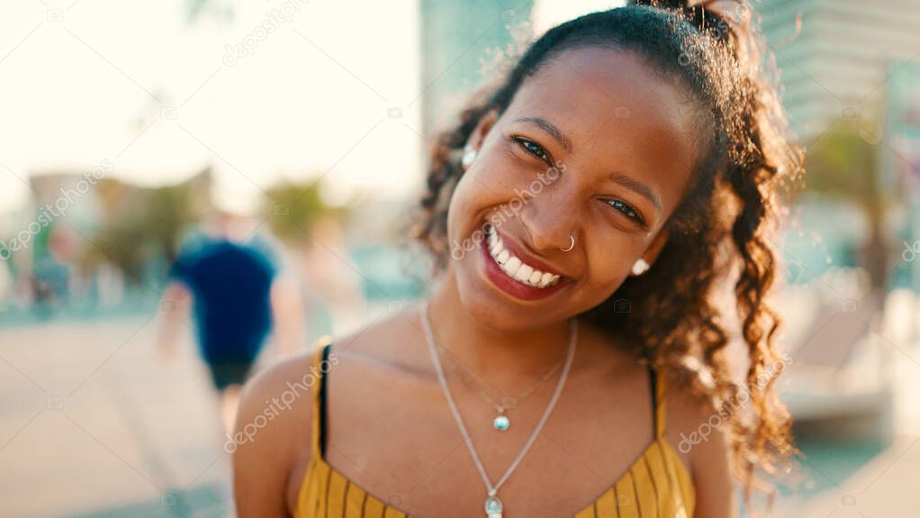 Closeup portrait of smiling woman with long curly hair on urban city background. Frontal close-up of happy girl fixing her hair and looking at the camera