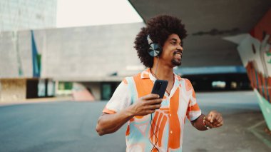 Tişört giyen, kulaklıkla müzik dinleyen ve şehir arka planında dans eden neşeli genç Afrikalı Amerikalı adamın yakın çekimi. Yaşam tarzı kavramı. 