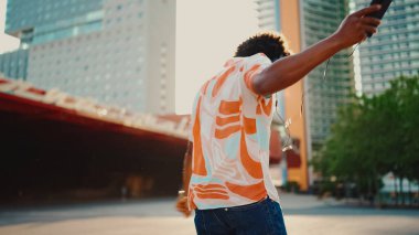 Tişört giyen, kulaklıkla müzik dinleyen ve şehir arka planında dans eden neşeli genç Afrikalı Amerikalı adamın yakın çekimi. Yaşam tarzı kavramı