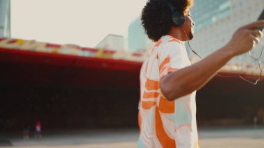 Tişört giyen, kulaklıkla müzik dinleyen ve şehir arka planında dans eden neşeli genç Afrikalı Amerikalı adamın yakın çekimi. Yaşam tarzı kavramı