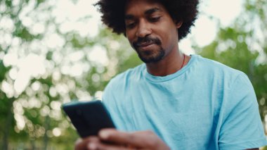Açık mavi tişörtlü, şehir bankında oturan ve akıllı telefonunu kullanan Afro-Amerikan bir adamın yakın plan portresi. Adam fotoğraflara, cep telefonundaki videolara bakıyor. yaşam tarzı konsepti.