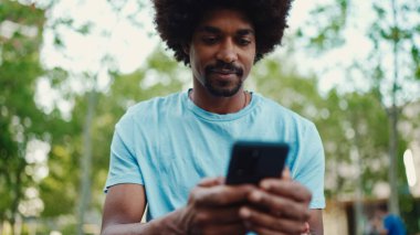 Açık mavi tişörtlü, şehir bankında oturan ve akıllı telefonunu kullanan Afro-Amerikan bir adamın yakın plan portresi. Adam fotoğraflara, cep telefonundaki videolara bakıyor. yaşam tarzı konsepti.
