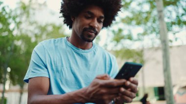 Açık mavi tişörtlü, şehir bankında oturan ve akıllı telefonunu kullanan Afro-Amerikan bir adamın yakın plan portresi. Adam fotoğraflara, cep telefonundaki videolara bakıyor. yaşam tarzı konsepti.