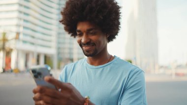Açık mavi tişörtlü genç Afro-Amerikan bir adamın akıllı telefonunu kullanarak yakın plan portresi. Adam fotoğraflara, cep telefonundaki videolara bakıyor.. 