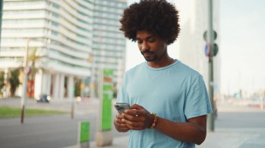Açık mavi tişörtlü genç Afro-Amerikan bir adamın akıllı telefonunu kullanarak yakın plan portresi. Adam fotoğraflara, cep telefonundaki videolara bakıyor.. 