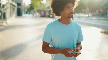 Açık mavi tişörtlü genç Afro-Amerikan bir adamın akıllı telefonunu kullanarak yakın plan portresi. Adam fotoğraflara, cep telefonundaki videolara bakıyor.. 