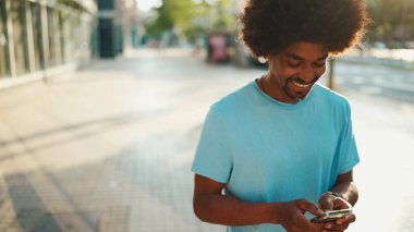 Açık mavi tişörtlü genç Afro-Amerikan bir adamın akıllı telefonunu kullanarak yakın plan portresi. Adam fotoğraflara, cep telefonundaki videolara bakıyor.. 