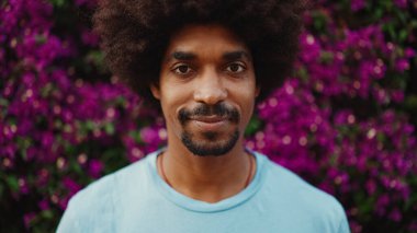 Genç Afro-Amerikalı bir adamın yakın plan portresi gözlerini açar kameraya bakar ve arka plan duvarında mor çiçeklerle kaplı bir gülümsemeyle durur. Kentsel yaşam. 