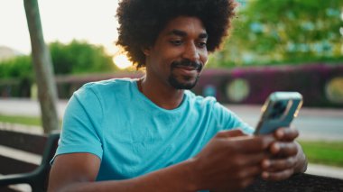 Mavi tişörtlü, parktaki bankta oturan ve hoparlörden konuşan Afro-Amerikan genç adamın yakın çekimi. Gülümseyen adam cep telefonundan sesli mesaj gönderiyor. yaşam tarzı konsepti. 