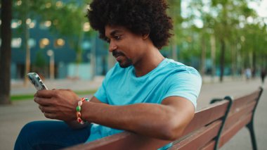Mavi tişörtlü, parktaki bankta oturan ve hoparlörden konuşan Afro-Amerikan genç adamın yakın çekimi. Gülümseyen adam cep telefonundan sesli mesaj gönderiyor. yaşam tarzı konsepti. 