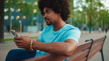 Mavi tişörtlü, parktaki bankta oturan ve hoparlörden konuşan Afro-Amerikan genç adamın yakın çekimi. Gülümseyen adam cep telefonundan sesli mesaj gönderiyor. yaşam tarzı konsepti. 