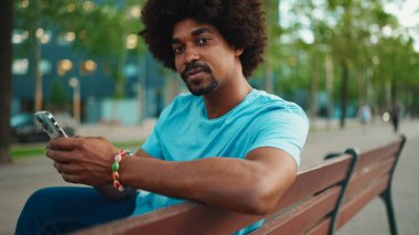 Mavi tişörtlü, parktaki bankta oturan ve hoparlörden konuşan Afro-Amerikan genç adamın yakın çekimi. Gülümseyen adam cep telefonundan sesli mesaj gönderiyor. yaşam tarzı konsepti. 