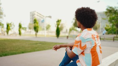 Parktaki bankta rahatça oturan, gömlek giyen Afro-Amerikalı genç adam başka tarafa bakıyor. Yaşam tarzı konsepti. 