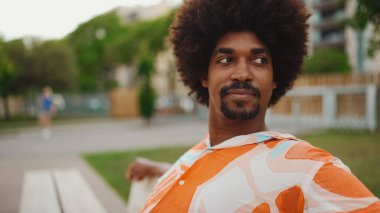 Parktaki bankta rahatça oturan, gömlek giyen Afro-Amerikalı genç adam başka tarafa bakıyor. Yaşam tarzı konsepti. 