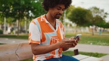 Parktaki bankta oturan Afro-Amerikan gömlekli genç adam akıllı telefonunu kullanıyor. Gülümseyen Afro-Amerikan bir adamın cep telefonundaki fotoğraf videosuna yakından bakması. Yaşam tarzı kavramı