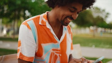 Parktaki bankta oturan Afro-Amerikan gömlekli genç adam akıllı telefonunu kullanıyor. Gülümseyen Afro-Amerikan bir adamın cep telefonundaki fotoğraf videosuna yakından bakması. Yaşam tarzı kavramı