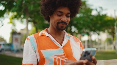 Parktaki bankta oturan Afro-Amerikan gömlekli genç adam akıllı telefonunu kullanıyor. Gülümseyen Afro-Amerikan bir adamın cep telefonundaki fotoğraf videosuna yakından bakması. Yaşam tarzı kavramı