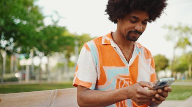 Parktaki bankta akıllı telefonuyla oturan, gömlek giyen Afro-Amerikan bir adamın yakın plan portresi. Gülümseyen adam cep telefonundan sesli mesaj gönderiyor.. 