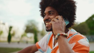 Parktaki bankta oturan Afro-Amerikan gömlekli genç adam akıllı telefonunu kullanıyor. Gülümseyen adam cep telefonuyla konuşuyor.. 