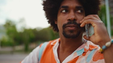 Parktaki bankta oturan Afro-Amerikan gömlekli genç adam akıllı telefonunu kullanıyor. Gülümseyen adam cep telefonuyla konuşuyor.. 