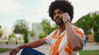 Parktaki bankta oturan Afro-Amerikan gömlekli genç adam akıllı telefonunu kullanıyor. Gülümseyen adam cep telefonuyla konuşuyor.. 