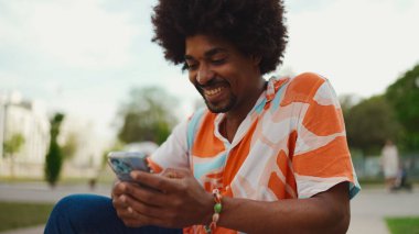 Parktaki bankta akıllı telefonuyla oturan, gömlek giyen Afro-Amerikan bir adamın yakın plan portresi. Yaşam tarzı kavramı. 