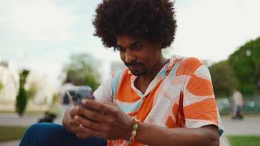 Parktaki bankta akıllı telefonuyla oturan, gömlek giyen Afro-Amerikan bir adamın yakın plan portresi. Yaşam tarzı kavramı. 